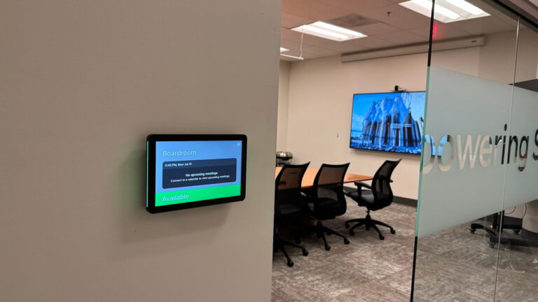 Room-scheduling panel showing a board room as available, with the glass-walled room and its display behind it.